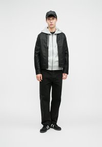 Giacca di pelle nera sopra un felpa grigia, abbinata a jeans neri e scarpe da ginnastica nere con strisce bianche. Il modello è in piedi contro uno sfondo semplice.