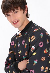 Giubbotto bomber nero con stampe colorate di alimenti come hamburger, ciambelle e gelato. Polsini e colletto a coste, texture imbottita.
