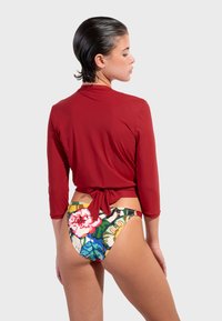 Crvena kupaća majica s tri četvrtine rukava, zavezana u struku; šareni badić donji dio s cvjetnim uzorkom koji uključuje zelene, plave i ružičaste nijanse.