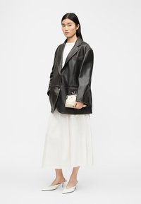 Mujer con blazer negro de cuero oversize, falda midi blanca, top blanco, tacones blancos puntiagudos y bolso de mano pequeño beige.