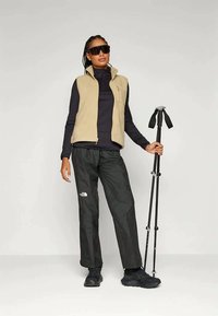 Μπεζ fleece γιλέκο πάνω από μαύρη μπλούζα με μακριά μανίκια, μαύρα αδιάβροχα παντελόνια, σκούρα μποτάκια πεζοπορίας και μαύρα γυαλιά ηλίου, με μπαστούνια πεζοπορίας.