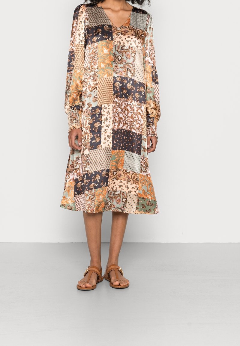 Mujer vistiendo un vestido midi de parches de manga larga con patrones florales y geométricos mezclados, combinado con sandalias marrones de punta abierta.