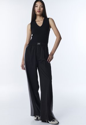 Jonge vrouw met een mouwloos zwart topje, zwarte wijde broek met grijze zijstrepen en zwart-witte schoenen, staand tegen een witte achtergrond.