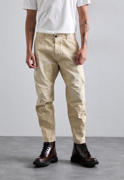 Persona che indossa pantaloni beige, stivali marroni scuri con lacci e una maglietta bianca, in piedi su un pavimento in cemento grigio di fronte a una parete semplice.
