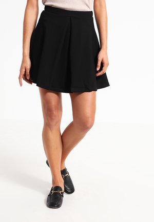 Woman wearing a black pleated mini skirt, beige sleeveless top, and black loafers with gold buckle details, standing with legs crossed.