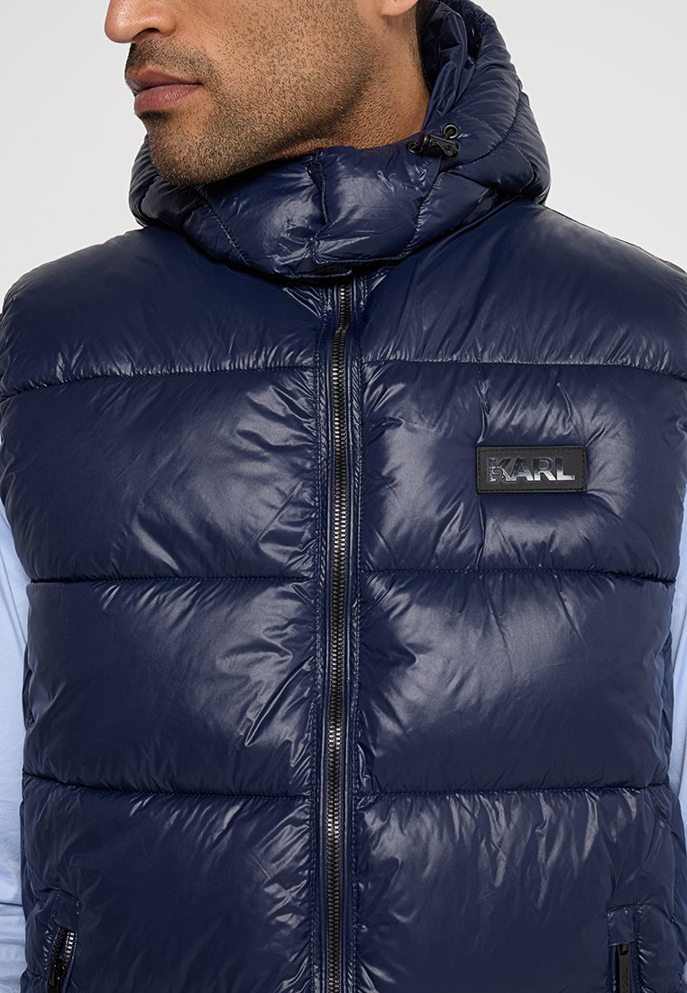 Gilet rembourré bleu marine avec col montant, fermeture éclair métallique et patch logo. Design matelassé en coussin, texture lisse et capuche ajustable.