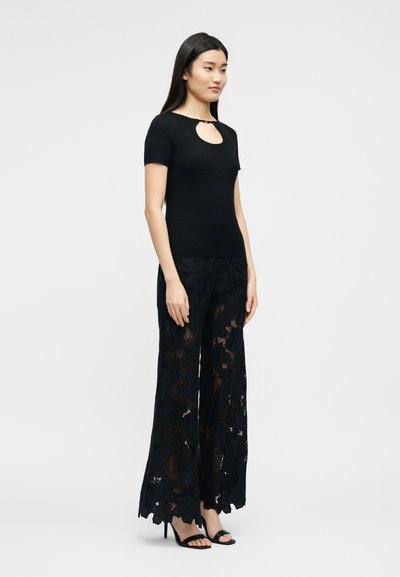 Γυναίκα που φοράει μαύρο μπλουζάκι με κοντά μανίκια και άνοιγμα στο μπροστινό μέρος, μαζί με φαρδιά μαύρα παντελόνια από δαντέλα με λουλουδένιο σχέδιο και μαύρα πέδιλα με ανοιχτά δάχτυλα, όρθια.