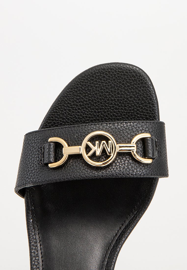 Sandale noire à glissière avec une sangle en cuir texturé et un accent logo doré circulaire. Semelle intérieure lisse avec une légère courbure pour le confort.