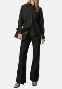 Camicia nera con bottoni e colletto classico, abbinata a jeans scuri svasati e una borsa nera con texture. Sandali neri chiusi.