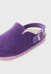 Violet filca klusņa ar mīkstu iekšpusi, rozā siksnu un atbilstošu zoli. Iespējas sānu sprādzes akcents un atvērta aizmugures dizains.