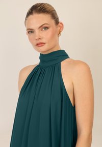 Teal halterneck kjole med samlet stof, med åben ryg og glat tekstur. Guld hoops øreringe fremhæver designet.