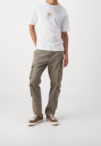 T-shirt bianca in cotone con grafica "Santorini", abbinata a pantaloni cargo beige con tasche laterali e vestibilità rilassata. Sneakers marroni completano il look.