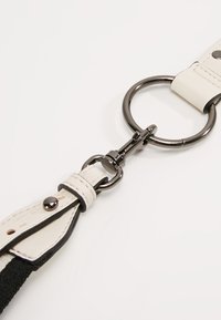 Fibbia portachiavi in pelle bianca con accenti neri, dotata di un anello metallico e di un gancio. Superficie strutturata con borchie. Design semplice e funzionale.