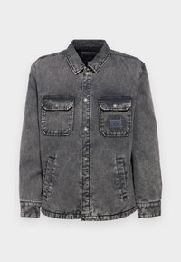 Chaqueta de mezclilla en gris desgastado con un cierre de botones en la parte frontal, dos bolsillos en el pecho y bolsillos laterales. Presenta una etiqueta de parche en contraste en el bolsillo izquierdo.