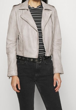 Žena v světle šedé semišové motorkářské bundě přes černobíle pruhované tričko, doplněné o černé džíny a pás s průchodkami.