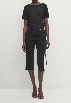 Personne portant un haut noir à manches courtes avec une fine ceinture, un pantalon noir court et des sandales rouges à lanières sur un fond clair uni.