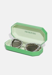 Swarovski slnečné okuliare v zelenom osmihrannom puzdre. Okuliare majú dizajn v tvare srdca, tmavé šošovky a rám zdobený kryštálmi.