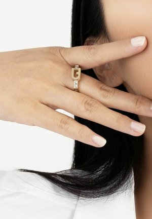 Gold ring featuring a stylized 'G' design and a band with clear gemstones, displayed on a finger with a neutral nail color.