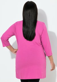 Kvinde med glat sort hår iført en lyserød langærmet tunika, stående med den ene hånd på hoften og vendt mod en hvid væg.