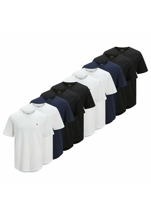 Giovane uomo con capelli corti che indossa una t-shirt bianca semplice con un piccolo logo sul petto e jeans blu, in piedi davanti a uno sfondo neutro.