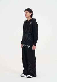 Zwarte hoodie met een zilver gesthebde vlamontwerp, ritssluiting en koorden; gecombineerd met zwarte wijde broek met gedurfde grafische tekst.