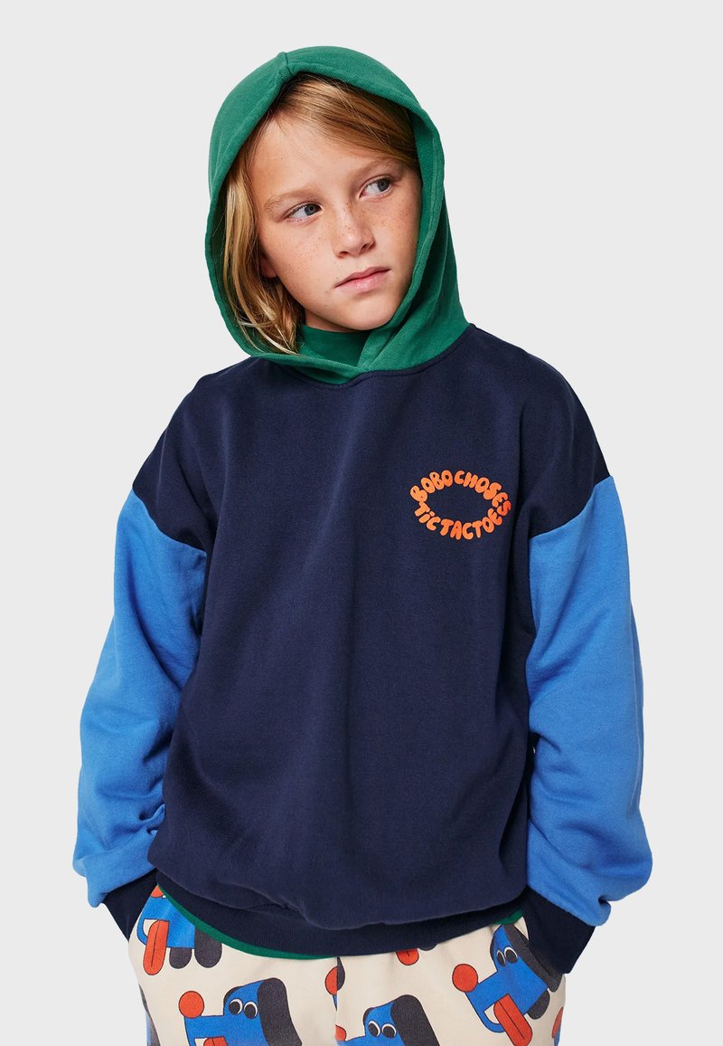 Felpa blu navy con cappuccio verde, maniche blu e logo arancione. Tessuto morbido, vestibilità comoda e polsini a costine. Pantaloni con motivo visibili.