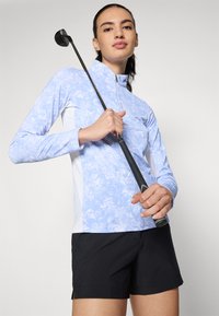 Kvinne i lys blå mønstret langermet skjorte og svarte shorts som holder en golfkølle oppreist mot en ensfarget bakgrunn.