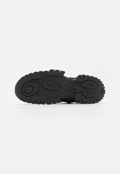Black rubber shoe sole with textured tread pattern, featuring raised grooves and a rounded heel for traction and stability.