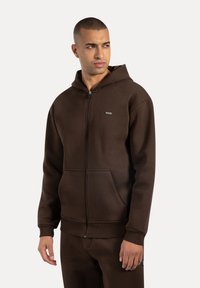 Sudadera con cremallera marrón hecha de tela suave, con capucha, bolsillos frontales, puños acanalados y un pequeño detalle de logo en el pecho.
