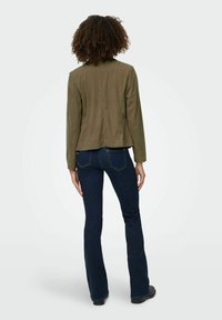 Olivgrüner Blazer mit gerader Silhouette, strukturiertem Stoff und Reverskragen, kombiniert mit dunkelblauen Schlagjeans und schwarzen Schuhen.
