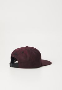 Bordeaux cap med flat brem, laget av teksturert stoff. Har svarte ventilasjonshull og en justerbar stropp bak.