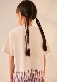 Világos bézs színű, rojtos szegélyű, rövidített pamut póló. Fonott haj hullik a hátoldalon, texturált kontrasztot teremtve.
