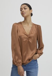 Blusa in satin marrone con scollo a V, dettagli arricciati sul petto e lunghe maniche puff con polsini elasticizzati. Indossata con jeans in denim blu.