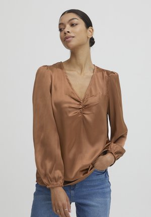 Braune Satinbluse mit V-Ausschnitt, gerafftem Detail an der Brust und langen Puffärmeln mit elastischen Bündchen. Kombiniert mit blauen Jeans.