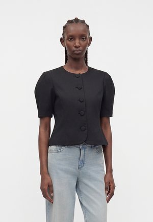 Γυναίκα φοράει μαύρο κοντομάνικο πουκάμισο με κουμπιά και φουσκωτούς ώμους και ανοιχτό μπλε ψηλόμεσο τζιν, στέκεται μπροστά σε λευκό φόντο.