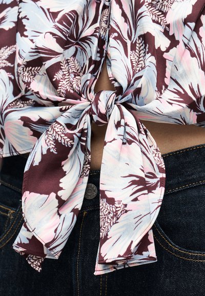 Κοντινό πλάνο πουκάμισου με λουλουδάτο μοτίβο δεμένου σε κόμπο πάνω από σκούρο τζιν με εμφανές κουμπί και ραφές.