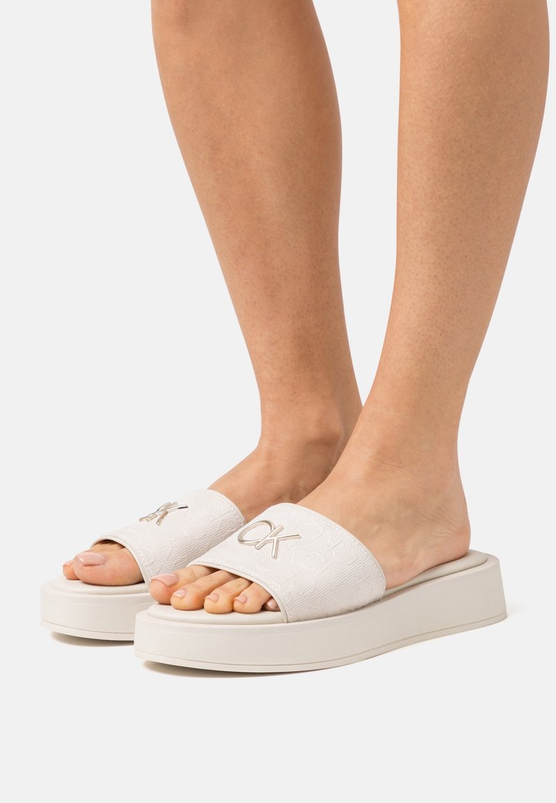 Pieds portant des sandales blanches à plateforme avec des brides texturées et un logo métallique "CK", présentés sur un fond clair uni.