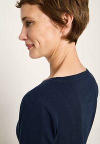 Frau mit kurzen braunen Haaren, die ein marineblaues Hemd trägt, seitlich gezeigt und leicht lächelnd vor einem hellen, einfarbigen Hintergrund.