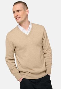 Pull beige à col en V en matière douce avec ourlet et poignets côtelés, superposé sur une chemise blanche à col, sur un fond uni.