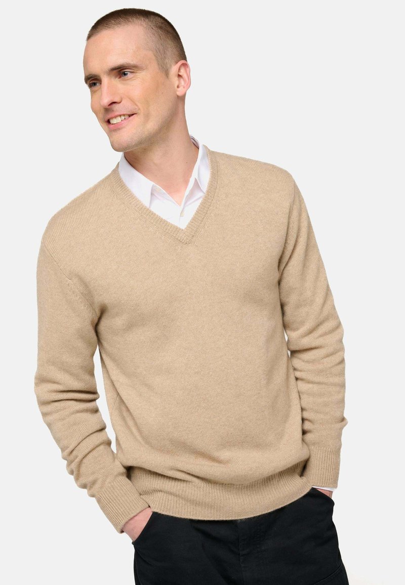 Pull beige à col en V en matière douce avec ourlet et poignets côtelés, superposé sur une chemise blanche à col, sur un fond uni.