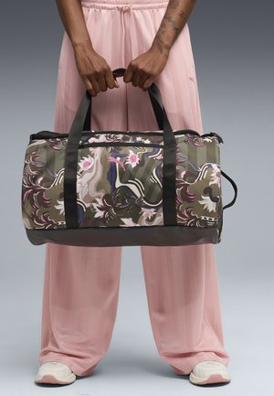 Person i lyserøde bukser og hvide sneakers holder en rejsetaske med blomsterprint og sorte håndtag mod en grå baggrund.