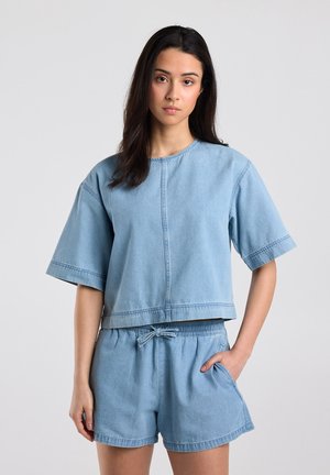 Frau mit langen dunklen Haaren, die ein hellblaues Denim-Kurzarmoberteil und passende Shorts mit Kordelzug trägt, stehend vor einem schlichten hellen Hintergrund.