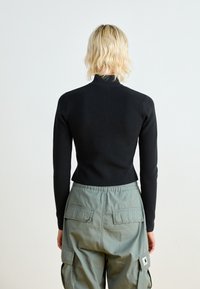 Person med axellångt blont hår bär en svart långärmad tröja och grågröna cargobyxor, stående mot en enkel ljus bakgrund.