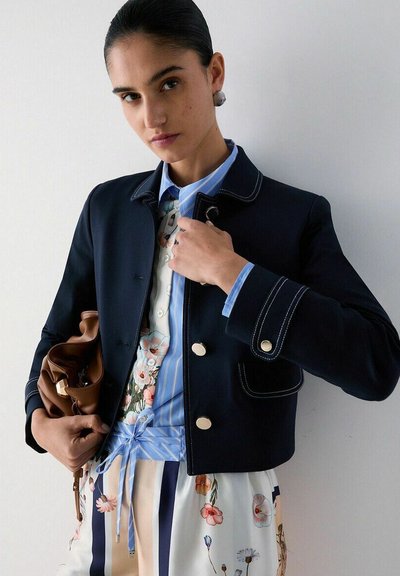 Femme portant une veste bleu marine sur une chemise rayée bleue et une jupe fleurie, tenant un sac en cuir marron sur un fond blanc uni.