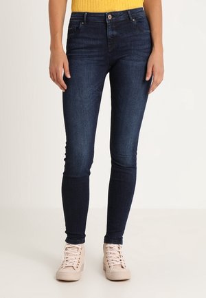 Jean slim bleu foncé porté avec des baskets rose pâle et un haut côtelé jaune, visible de la taille aux pieds sur un fond uni.