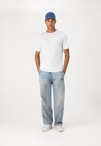 Λευκό κοντομάνικο μπλουζάκι, ανοιχτό μπλε φαρδύ τζιν με κόκκινο λογότυπο και λευκά αθλητικά παπούτσια. Το μοντέλο φορά επίσης ένα μπλε καπέλο.