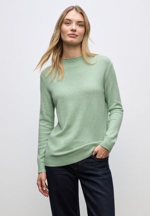 Lichtgroene gebreide trui met een ronde hals en geribbelde boorden. Gepaard met donkerblauwe jeans, weergegeven tegen een neutrale achtergrond.