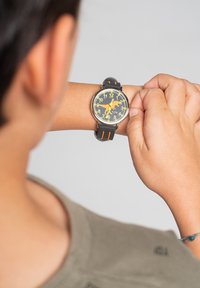 Montre noire avec un cadran rond, accents jaunes et oranges, chiffres audacieux et un bracelet en silicone texturé, présente un élément de design ludique.