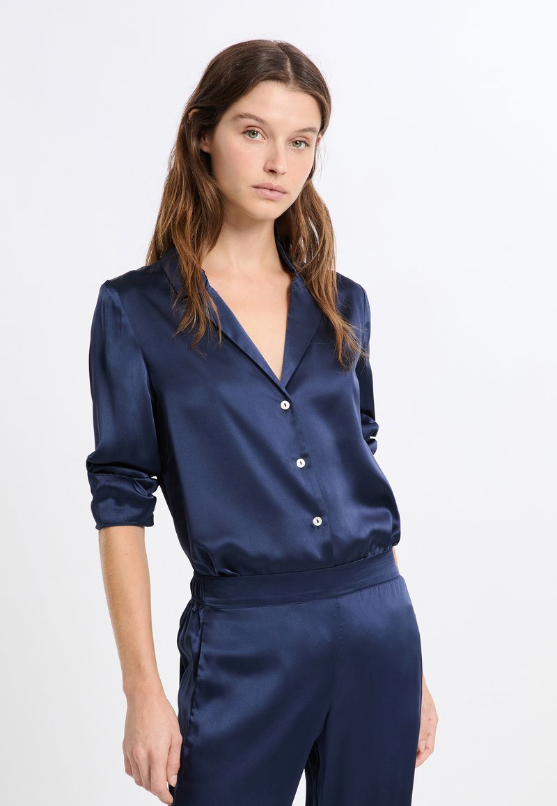 Blusa de satén azul marino con mangas largas, cuello con muesca y cierre de botones. Textura suave con un ligero brillo. Combinada con pantalones a juego.