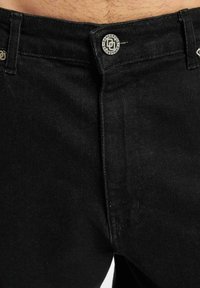 Schwarze Denim-Jeans mit glatter Textur, klassischem Fünf-Taschen-Design und Metallknopf mit "Dangerous"-Branding am Bund.